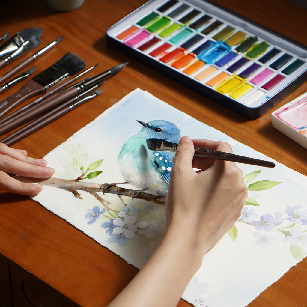 Person painting a blue bird on a branch with watercolors and brushes.